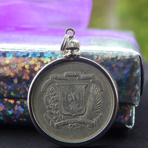 May include: Silver pendant featuring the coat of arms of the Dominican Republic. The coat of arms is a shield with a cross, a book, and a palm tree. The inscription around the shield reads "Dios, Patria, Libertad" and "República Dominicana".