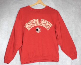 May include: A red long-sleeve crewneck sweatshirt featuring the text FLORIDA STATE in an arched gold font with a white outline. Below the text is a circular logo depicting a profile of a person with a feather in their hair.