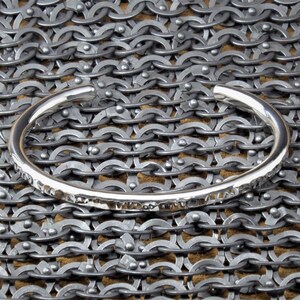 Peut inclure: Un bracelet manchette en argent avec une texture martel&eacute;e. Le bracelet est pr&eacute;sent&eacute; sur un fond de cotte de mailles, mettant en valeur sa surface r&eacute;fl&eacute;chissante et son design simple. Le bracelet ouvert est un bijou minimaliste.