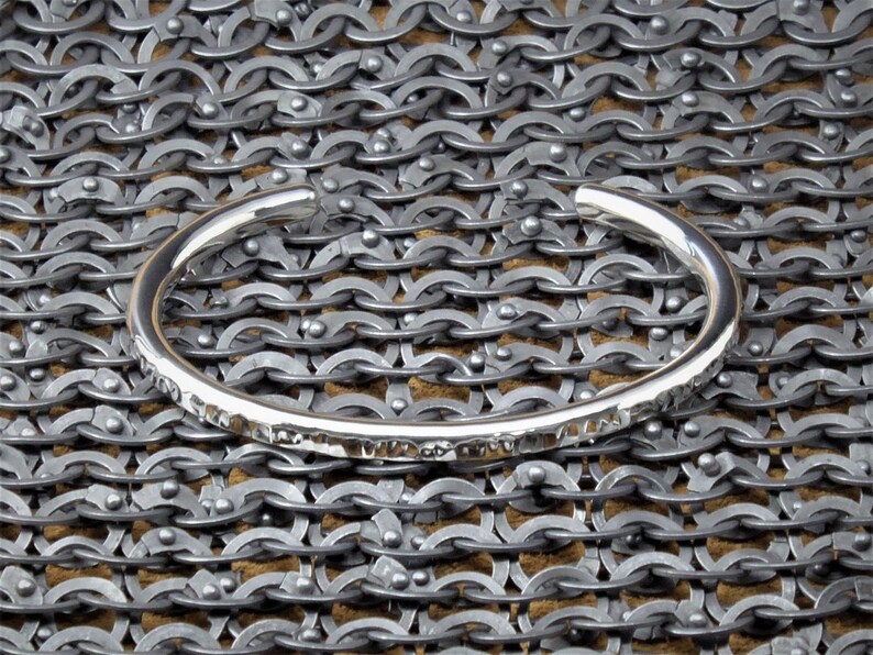 Peut inclure: Un bracelet manchette en argent avec une texture martel&eacute;e. Le bracelet est pr&eacute;sent&eacute; sur un fond de cotte de mailles, mettant en valeur sa surface r&eacute;fl&eacute;chissante et son design simple. Le bracelet ouvert est un bijou minimaliste.