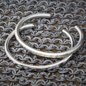 Peut inclure: Deux bracelets de manchette argent&eacute;s. L'un est lisse, l'autre textur&eacute;. Les deux sont ouverts et reposent sur une surface ressemblant &agrave; une cotte de mailles.