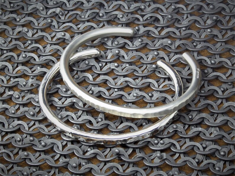 Peut inclure: Deux bracelets de manchette argent&eacute;s. L'un est lisse, l'autre textur&eacute;. Les deux sont ouverts et reposent sur une surface ressemblant &agrave; une cotte de mailles.