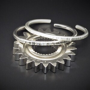 Peut inclure: Trois bracelets argent&eacute;s avec des textures martel&eacute;es reposent sur un engrenage argent&eacute;. Les bracelets ouverts ont un design simple et &eacute;l&eacute;gant. L'engrenage a plusieurs dents et est pos&eacute; sur un fond sombre.