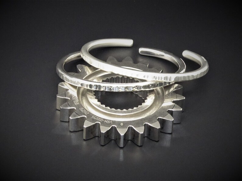 Peut inclure: Trois bracelets argent&eacute;s avec des textures martel&eacute;es reposent sur un engrenage argent&eacute;. Les bracelets ouverts ont un design simple et &eacute;l&eacute;gant. L'engrenage a plusieurs dents et est pos&eacute; sur un fond sombre.