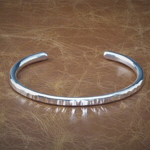 Peut inclure: Un bracelet manchette en argent avec une texture martel&eacute;e. Le bracelet ouvert est de conception simple et &eacute;l&eacute;gante. Le m&eacute;tal refl&egrave;te la lumi&egrave;re, cr&eacute;ant des reflets et des ombres. Le bracelet est pr&eacute;sent&eacute; sur une surface marron.