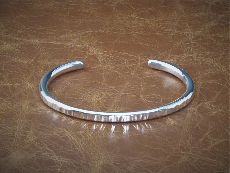 Peut inclure: Un bracelet manchette en argent avec une texture martel&eacute;e. Le bracelet ouvert est de conception simple et &eacute;l&eacute;gante. Le m&eacute;tal refl&egrave;te la lumi&egrave;re, cr&eacute;ant des reflets et des ombres. Le bracelet est pr&eacute;sent&eacute; sur une surface marron.