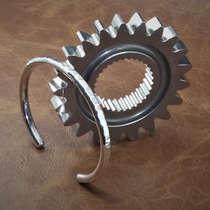 Peut inclure: Un bracelet manchette ouvert de couleur argent&eacute;e avec une texture martel&eacute;e est plac&eacute; devant un grand engrenage argent&eacute;. L'engrenage a un centre circulaire avec des dents internes et une s&eacute;rie de dents externes. Le fond est une surface brune textur&eacute;e.