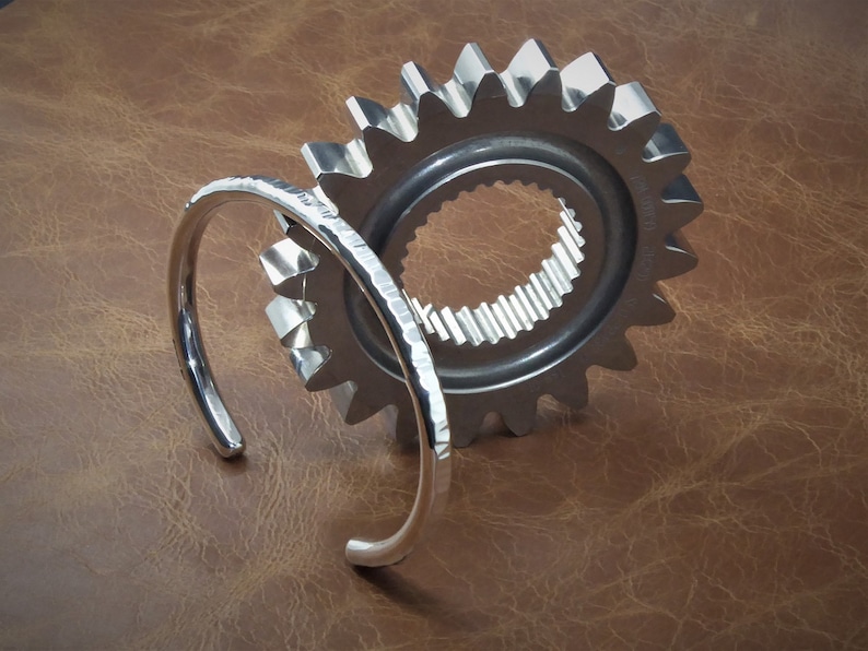 Peut inclure: Un bracelet manchette ouvert de couleur argent&eacute;e avec une texture martel&eacute;e est plac&eacute; devant un grand engrenage argent&eacute;. L'engrenage a un centre circulaire avec des dents internes et une s&eacute;rie de dents externes. Le fond est une surface brune textur&eacute;e.