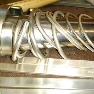 Peut inclure: Gros plan sur des bracelets et des bandes en m&eacute;tal argent&eacute;, probablement pour la fabrication de bijoux. Les bracelets sont enroul&eacute;s autour d'un cylindre m&eacute;tallique. Les bandes, avec du texte estamp&eacute;, sont pos&eacute;es sur une surface brune.