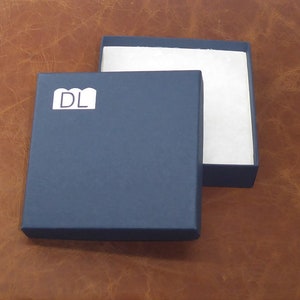 Peut inclure: Une bo&icirc;te carr&eacute;e bleu marine avec un int&eacute;rieur blanc et le couvercle l&eacute;g&egrave;rement entrouvert. La bo&icirc;te porte une &eacute;tiquette blanche avec les lettres "DL" sur le devant. La bo&icirc;te est pos&eacute;e sur une surface en cuir marron.