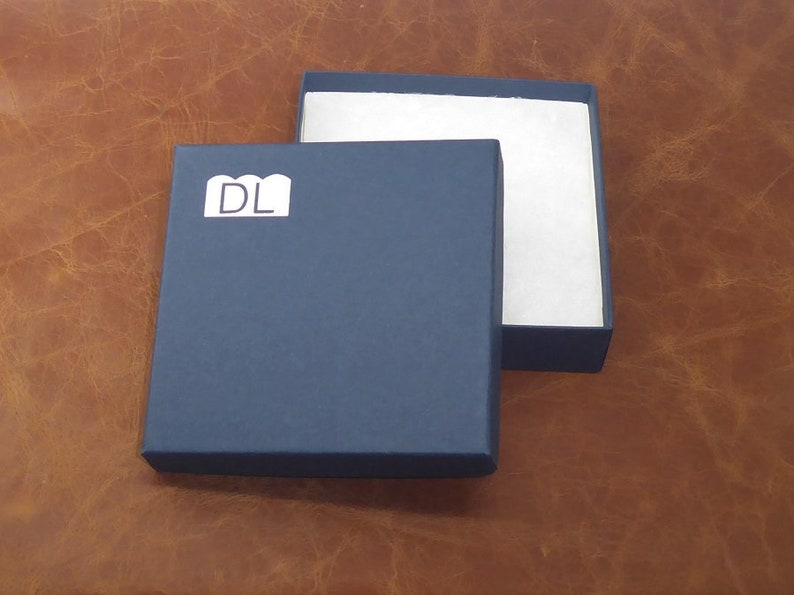 Peut inclure: Une bo&icirc;te carr&eacute;e bleu marine avec un int&eacute;rieur blanc et le couvercle l&eacute;g&egrave;rement entrouvert. La bo&icirc;te porte une &eacute;tiquette blanche avec les lettres "DL" sur le devant. La bo&icirc;te est pos&eacute;e sur une surface en cuir marron.
