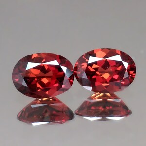 May include: Two oval-shaped red gemstones with a faceted cut. The gemstones are set against a white background.