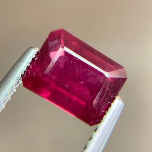 May include: A faceted, rectangular ruby gemstone held by silver tweezers. The ruby is a deep, saturated red colour with visible internal inclusions. The gemstone's cut features multiple facets that reflect light.