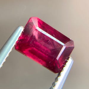以下が含まれることがあります： 銀色のピンセットで保持された、長方形のファセットルビーの宝石。 ルビーは、内部のインクルージョンが見える、深く飽和した赤色をしています。 宝石のカットは、そのシャープなエッジと研磨された表面を際立たせています。
