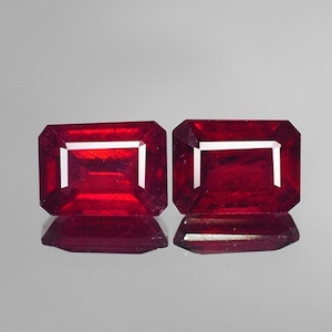 May include: Two deep red rectangular gemstones with a faceted cut. The gemstones are set in a clear, faceted setting.
