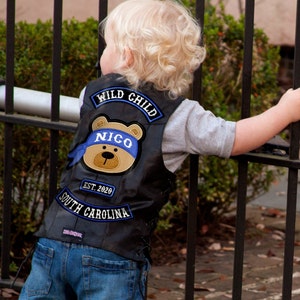 May include: A black leather vest with a blue and white bear patch that says "Nico" and a blue patch that says "Wild Child". The vest also has a blue patch that says "Est. 2020 South Carolina".