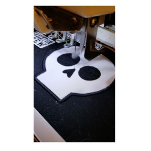 May include: Close-up of an embroidery machine stitching a white skull patch onto black fabric. The skull has black embroidered details and a black border. The machine's needle is in action, creating the design.