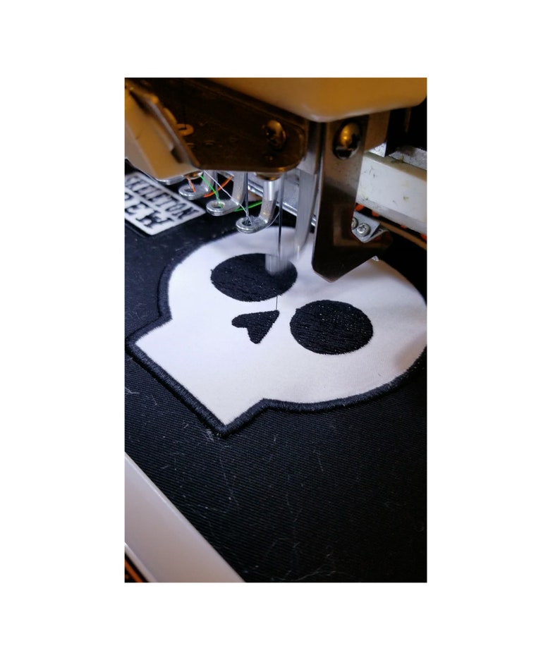 May include: Close-up of an embroidery machine stitching a white skull patch onto black fabric. The skull has black embroidered details and a black border. The machine's needle is in action, creating the design.