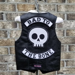 Pode incluir: Colete de couro preto com um patch de caveira branca e as palavras "BAD TO THE BONE" e "Kid Biker" em letras brancas. O colete está pendurado em um cabide contra uma parede de tijolos.