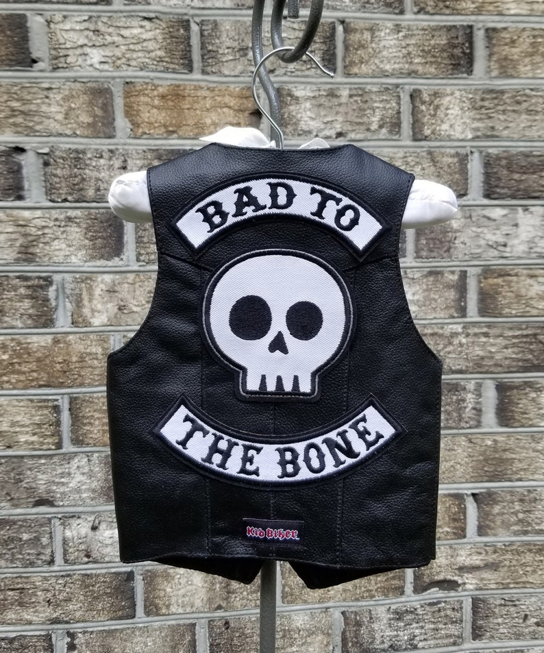 May include: Black leather vest with a white skull patch and the words "BAD TO THE BONE" and "Kid Biker" in white lettering. The vest is hanging on a hanger against a brick wall.