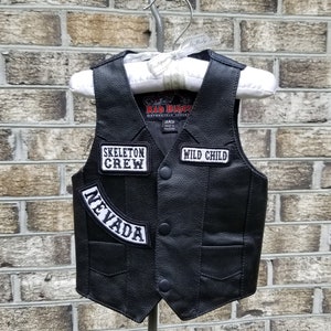 May include: Black leather vest with three patches: "SKELETON CREW", "WILD CHILD", and "NEVADA". The vest has a classic biker style with a button-up front and two front pockets. The vest is hanging on a white hanger.