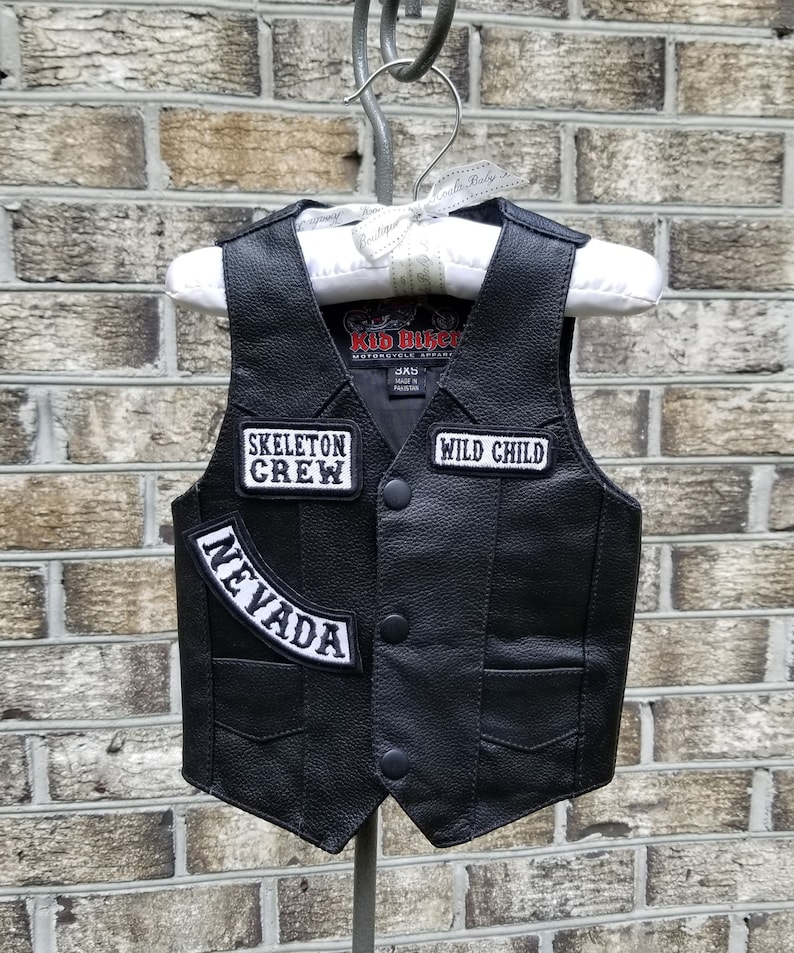 May include: Black leather vest with three patches: "SKELETON CREW", "WILD CHILD", and "NEVADA". The vest has a classic biker style with a button-up front and two front pockets. The vest is hanging on a white hanger.