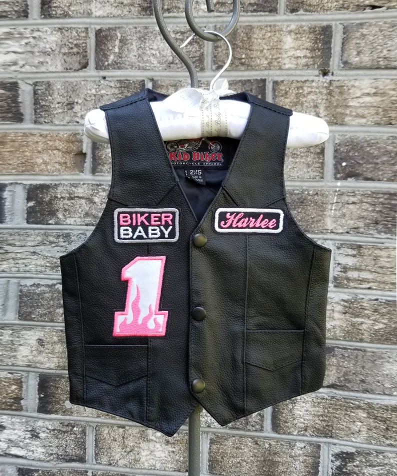 May include: Black leather vest with pink patches that say "Biker Baby", "Harlee", and "1" with a flame design. The vest has a Kid Biker Motorcycle Apparel label and size tag.
