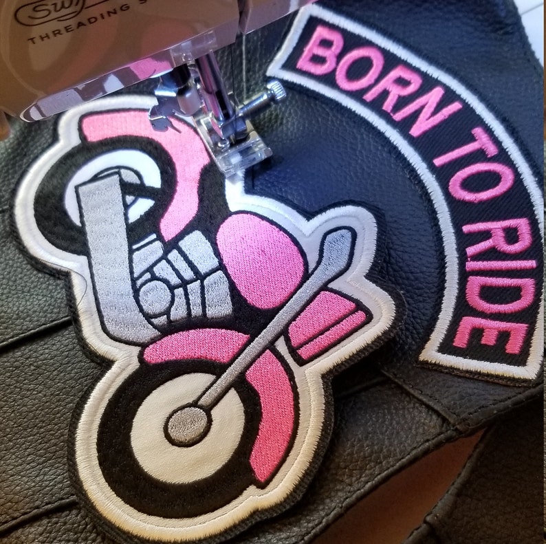 May include: Pink and white embroidered motorcycle patch with the words "Born to Ride" in a pink arc above the motorcycle. The patch is being sewn onto a black leather jacket.