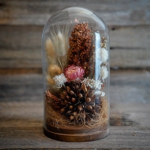 May include: A glass dome with a wooden base containing dried flowers and a pine cone. The flowers are white, pink, and brown. The pine cone is brown and has a natural texture.