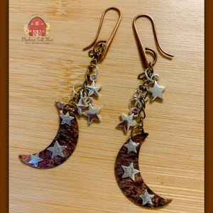 May include: A pair of handcrafted earrings featuring a crescent moon design with silver star accents. The earrings have copper-colored ear wires and chains, with a rustic, textured finish on the moon pendants. The earrings are displayed on a wooden surface.