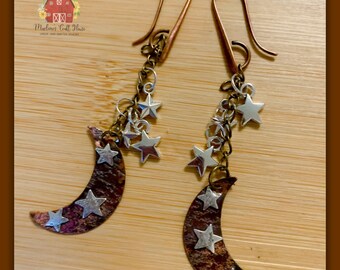 May include: A pair of handcrafted earrings featuring a crescent moon design with silver star accents. The earrings have copper-colored ear wires and chains, with a rustic, textured finish on the moon pendants. The earrings are displayed on a wooden surface.