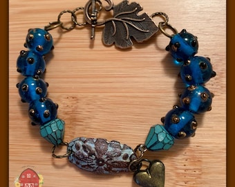 Pode incluir: Um bracelete de contas de vidro azul com detalhes dourados e um fecho de folha de bronze. O bracelete também tem uma pedra preciosa turquesa e um fecho de coração de bronze.