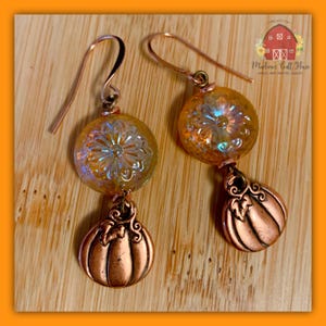 Puede incluir: Un par de pendientes colgantes con una cuenta de vidrio redonda de color ámbar iridiscente con un patrón floral, suspendida sobre un dije de calabaza de metal en tono cobre. Los pendientes están sujetos a ganchos de metal color cobre.