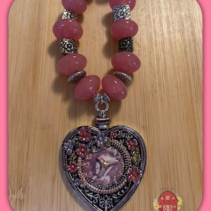 Peut inclure: Un collier de perles roses avec un pendentif en forme de cœur. Le pendentif est de couleur argentée avec un motif de cadran d'horloge et des accents floraux. Le collier comporte de grosses perles roses et des entretoises décoratives argentées.