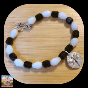 Op de afbeelding: Een armband met afwisselend zwarte en witte kralen. De armband heeft een zilveren toggle sluiting en een zilveren bedel met een libellen ontwerp. De kralen zijn facet geslepen en hebben een glanzende afwerking.