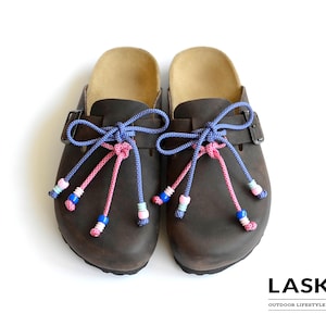 May include: A pair of dark brown slip-on clogs featuring adjustable side buckles and decorative blue and pink rope laces tied in bows. The laces are adorned with small, colorful beads at the ends. The text LASKO OUTDOOR LIFESTYLE DESIGN is visible.