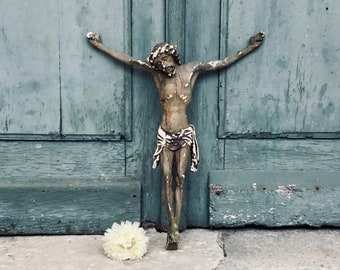 Könnte beinhalten: Eine bronzene Kruzifix-Statue mit ausgestreckten Armen steht auf einer Steinstufe vor einer verwitterten türkisblauen Tür. Eine kleine weiße Blume liegt in der Nähe des Sockels der Statue.