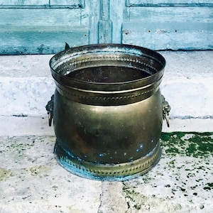 Puede incluir: Una jardinera de latón vintage con una parte superior ancha y abierta y asas decorativas con cabeza de león. La jardinera tiene una pátina desgastada, lo que sugiere edad y uso. Se encuentra sobre una superficie de piedra frente a una puerta azul claro desgastada.