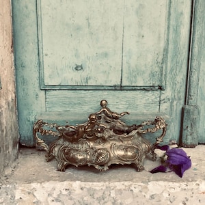 Może przedstawiać: Antyczny, złoty pojemnik dekoracyjny z bogatymi zdobieniami, przedstawiający postacie i ornamenty. Obok leży fioletowy kwiat. Pojemnik ustawiony jest na tle zwietrzałych, jasnoniebieskich drewnianych drzwi.