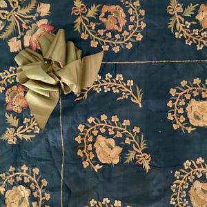 Può includere: Sezione di tessuto blu scuro con motivi floreali ricamati in beige, rosa e verde. Un grande fiocco di raso verde oliva è attaccato. Il ricamo presenta dettagliati disegni di fiori e foglie. Il tessuto è parte di un pezzo più grande.