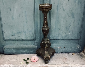 May include: An antique, ornate metal candle holder with a wide, fluted top and a detailed base. The candle holder is dark with a weathered appearance. A pink rose with green leaves rests on the stone step in front of a blue door.