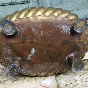K&ouml;nnte beinhalten: Antiker Messing-Pflanztopf mit einem seilartigen Rand und vier dekorativen F&uuml;&szlig;en. Der ovale Pflanztopf hat eine angelaufene Patina, die Gebrauchsspuren aufweist. Ein kleiner Griff ist auf einer Seite sichtbar. Der Pflanztopf steht auf einer Steinoberfl&auml;che.