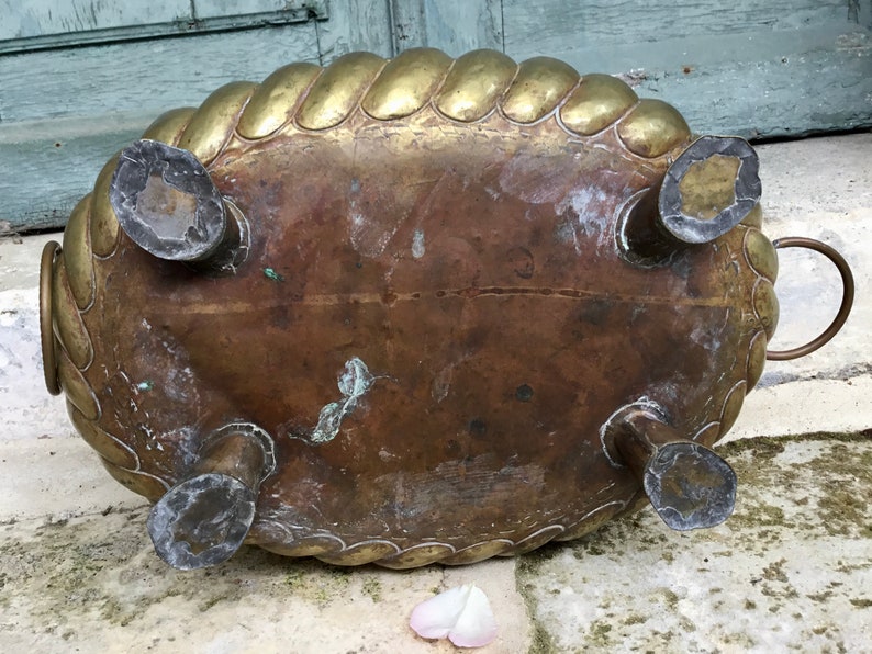 K&ouml;nnte beinhalten: Antiker Messing-Pflanztopf mit einem seilartigen Rand und vier dekorativen F&uuml;&szlig;en. Der ovale Pflanztopf hat eine angelaufene Patina, die Gebrauchsspuren aufweist. Ein kleiner Griff ist auf einer Seite sichtbar. Der Pflanztopf steht auf einer Steinoberfl&auml;che.