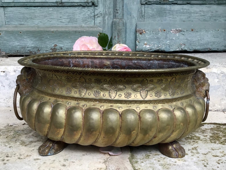 K&ouml;nnte beinhalten: Antiker Messing-Pflanzk&uuml;bel mit L&ouml;wenkopfgriffen und Klauenf&uuml;&szlig;en. Der Pflanzk&uuml;bel hat ein dekoratives Blumen- und geometrisches Muster. Einige rosa Rosen sind im Inneren des Pflanzk&uuml;bels platziert.