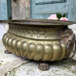 K&ouml;nnte beinhalten: Antiker Messing-Blumentopf mit floralen Details und L&ouml;wenkopf. Der Pflanztopf hat eine breite, flache Schalenform mit dekorativem Rand. Die Seiten sind gerippt und der Pflanztopf steht auf drei Klauenf&uuml;&szlig;en. Eine rosa Rose liegt im Inneren des Pflanztopfs.