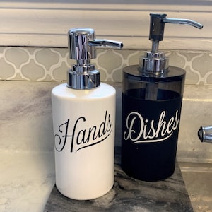 May include: Two white ceramic soap dispensers with black lettering. The dispenser on the left says "Hands" and the dispenser on the right says "Dishes".