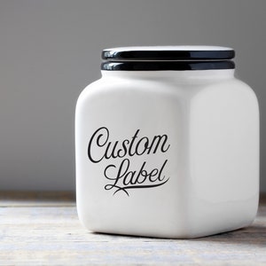 May include: White ceramic jar with a black lid and a black cursive label that says "Custom Label".