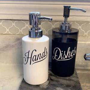 May include: Two white ceramic soap dispensers with black lettering. The dispenser on the left says "Hands" and the dispenser on the right says "Dishes".