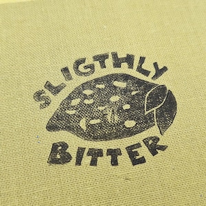 May include: Close-up of a block print on a textured, light green surface. The print features a black illustration of a lemon with the words "SLIGHTLY BITTER" above and below the lemon. The artist's name and "block prints" are at the bottom.