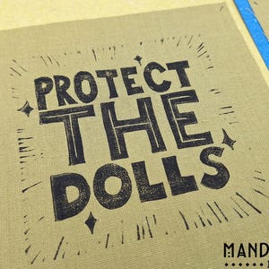 Op de afbeelding: Close-up van verschillende vierkante blokprints met de tekst "PROTECT THE DOLLS" in vette zwarte letters. De prints staan op tan papier met een getextureerd uiterlijk, de achtergrond is wit en blauw.